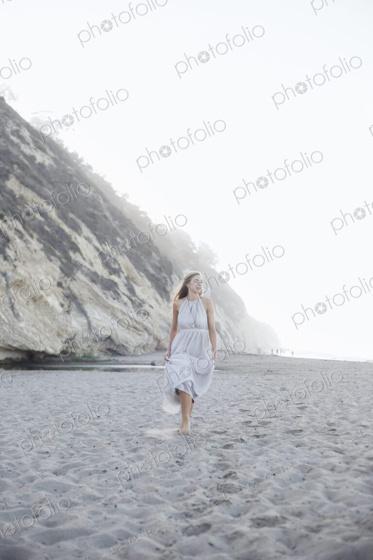Blond woman walking on a sandy beach near a cliff.
