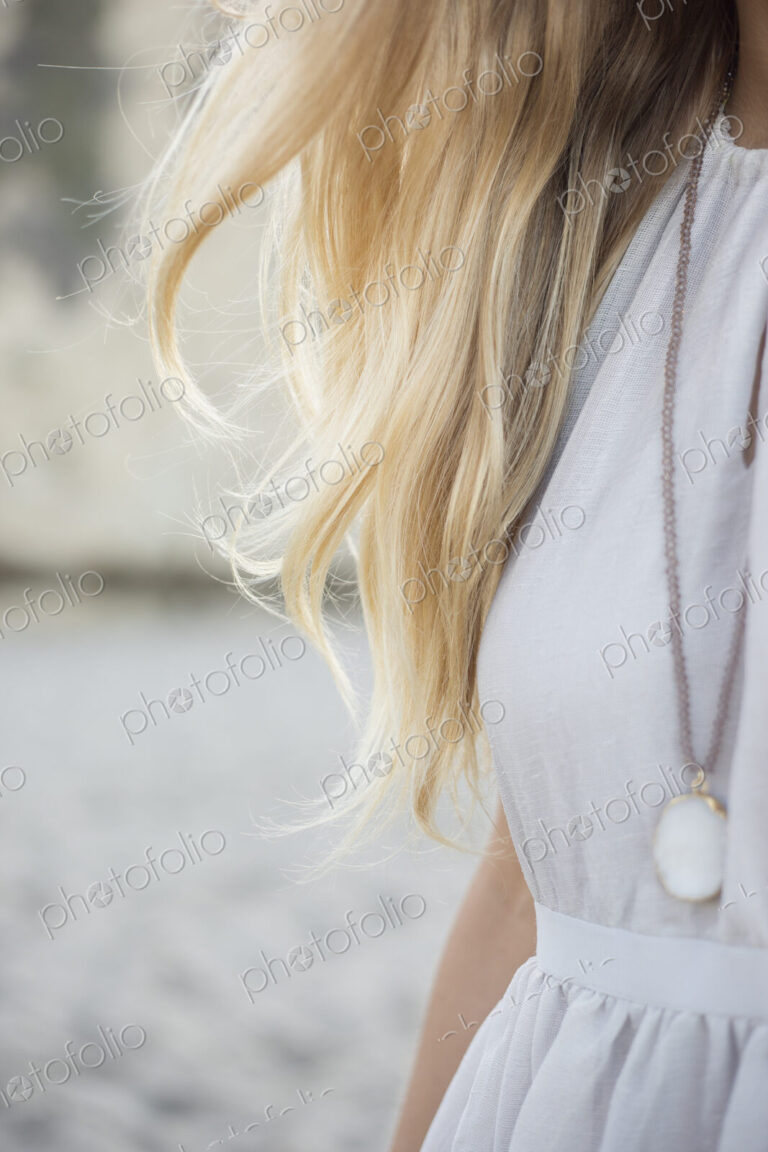Blond woman wearing a dress and necklace on a sandy beach.