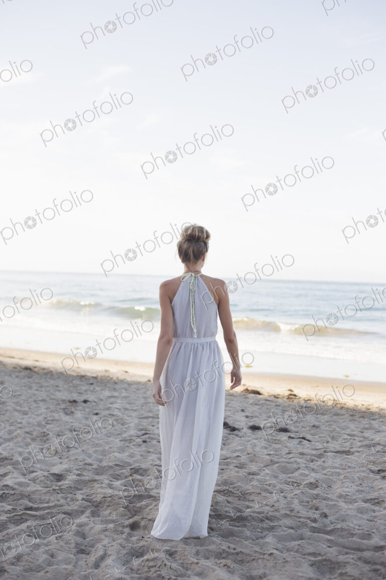 Blond woman wearing a long dress standing on a sandy beach.