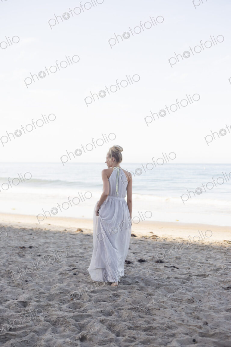 Blond woman wearing a long dress standing on a sandy beach.