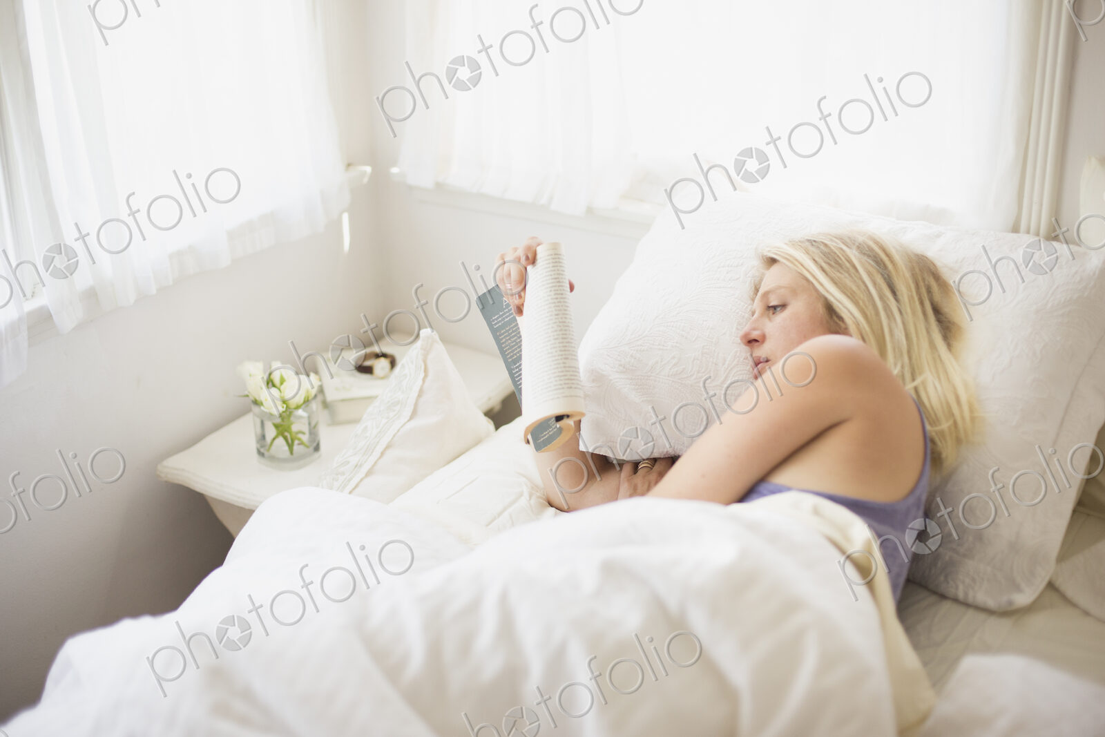 Blonde woman reading in a bed with white linen.
