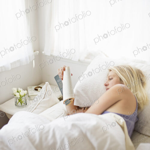 Blonde woman reading in a bed with white linen.