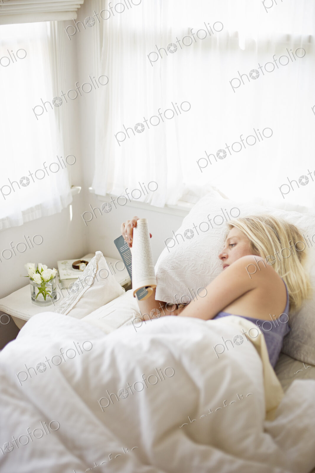 Blonde woman reading in a bed with white linen.