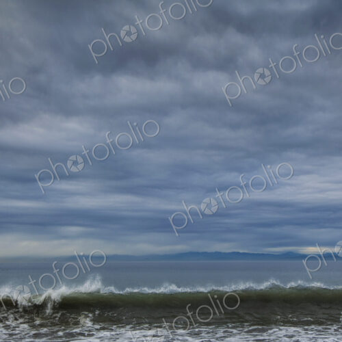 Breaking waves and view across the ocean from the coast under a cloudy sky.