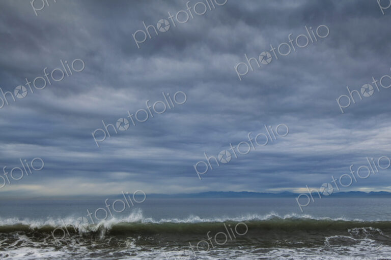 Breaking waves and view across the ocean from the coast under a cloudy sky.