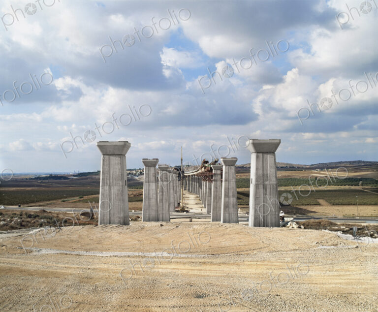 Bridge under construction, pillars and elevated road across an arid landscape.