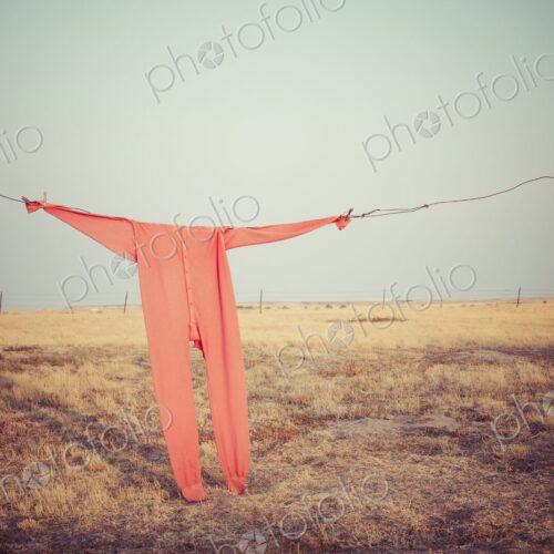 Bright red long johns hang from a wire clothesline between fence posts on a wide South…