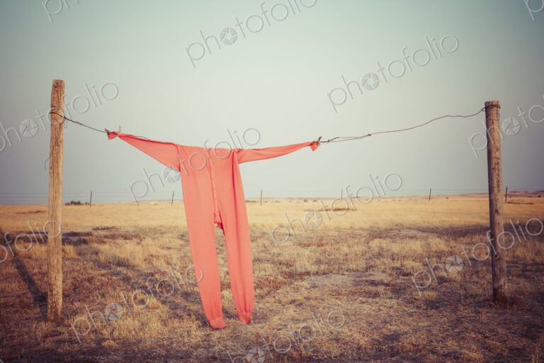 Bright red long johns hang from a wire clothesline between fence posts on a wide South…