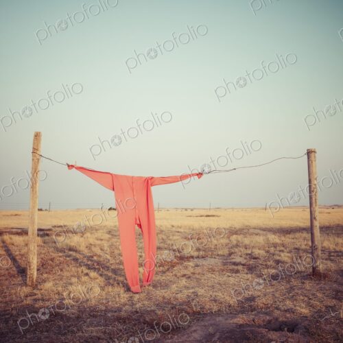 Bright red long johns hang from a wire clothesline between fence posts on a wide South…