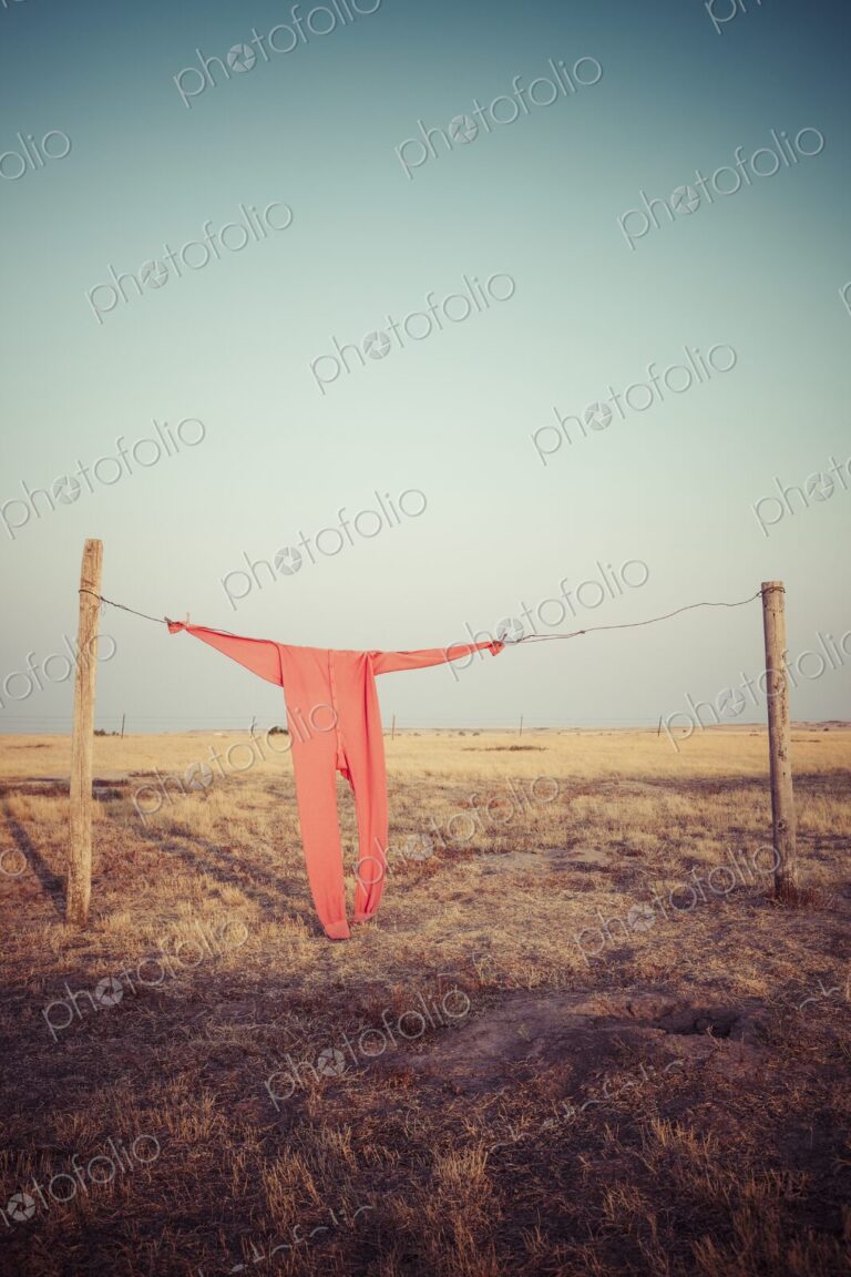 Bright red long johns hang from a wire clothesline between fence posts on a wide South…