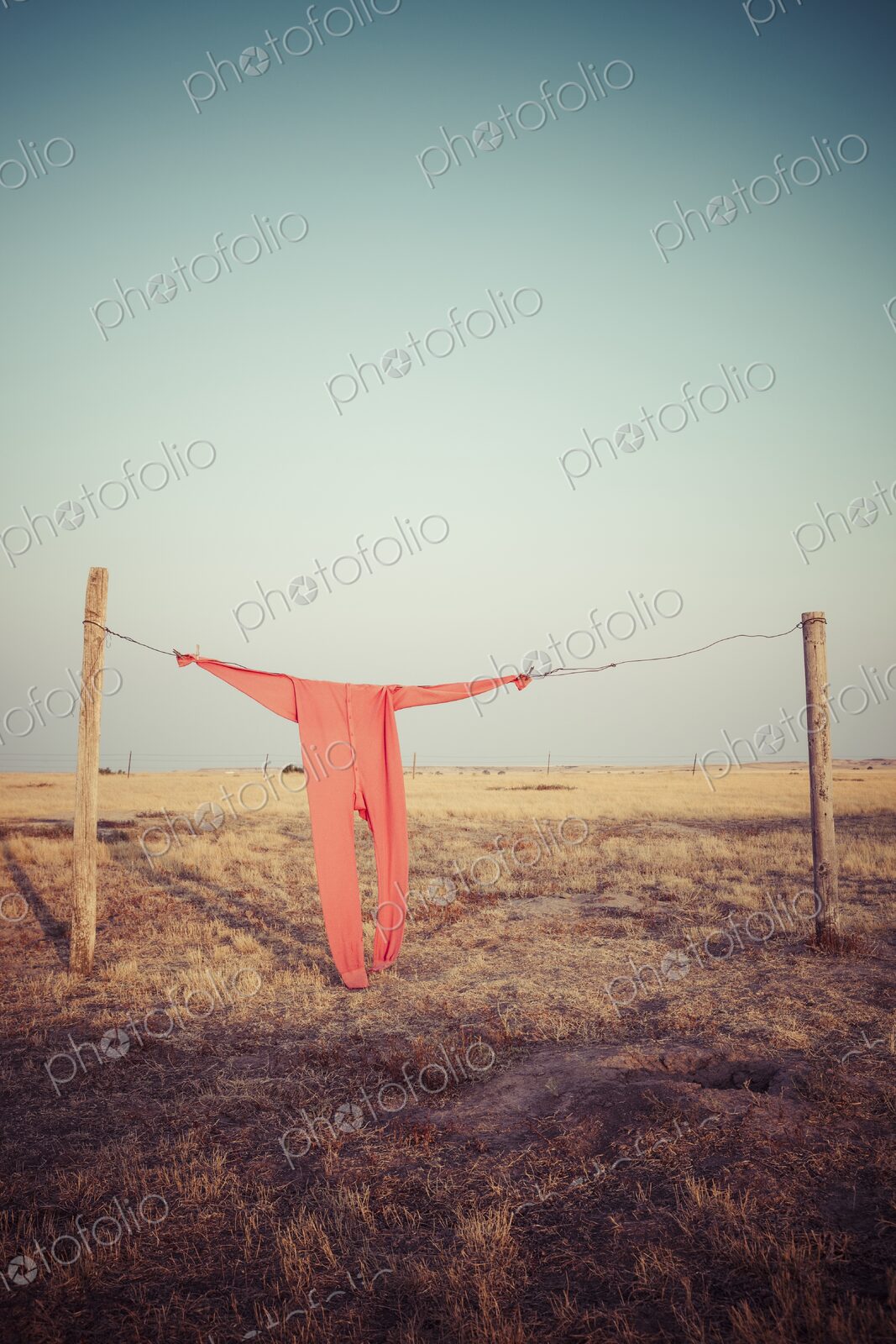 Bright red long johns hang from a wire clothesline between fence posts on a wide South…