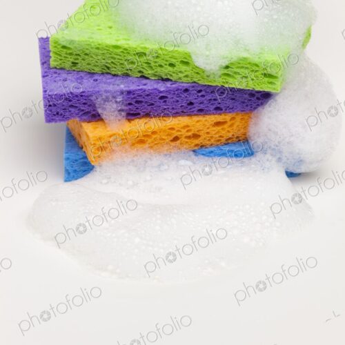 Brightly colored household sponges stacked and covered in foamy soap bubbles against a…