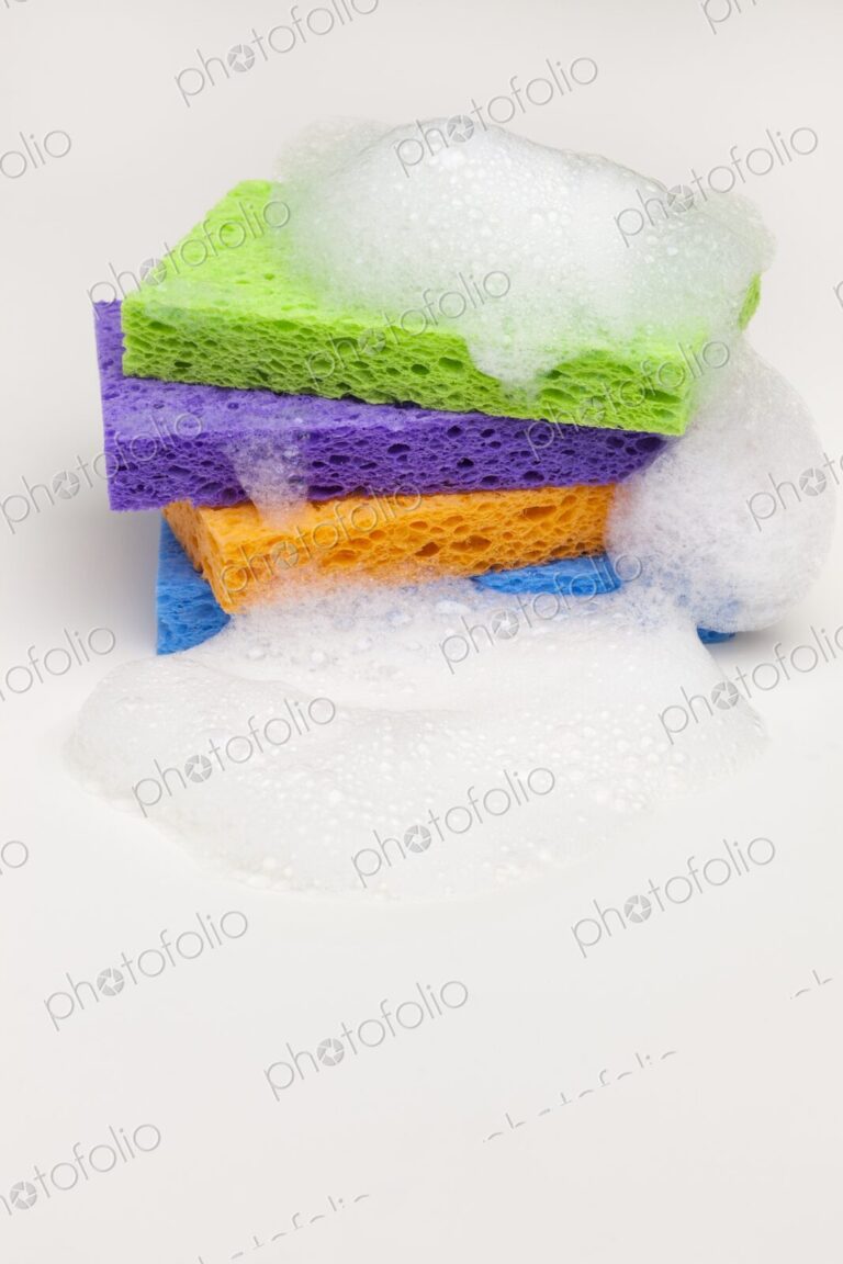 Brightly colored household sponges stacked and covered in foamy soap bubbles against a…