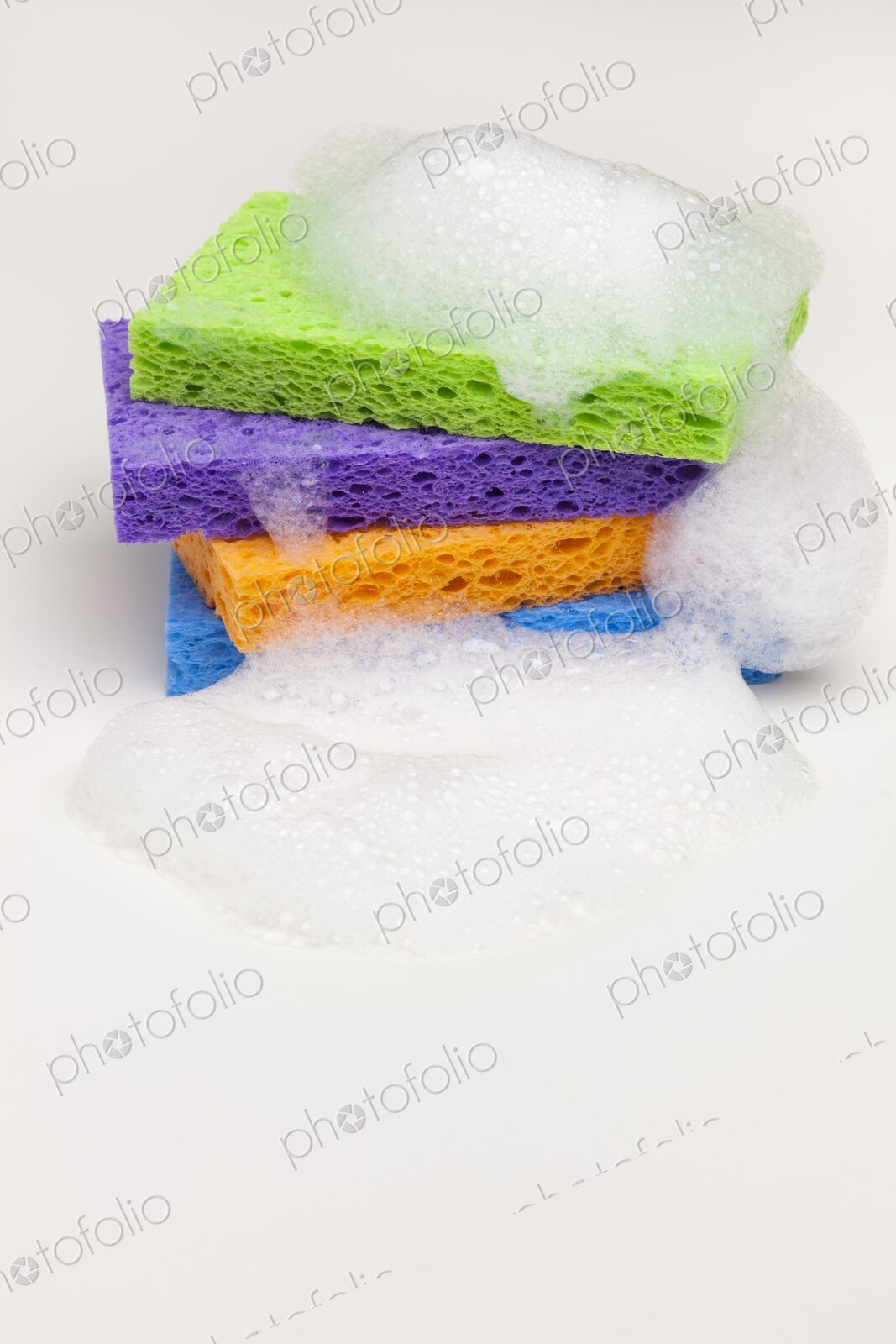 Brightly colored household sponges stacked and covered in foamy soap bubbles against a…