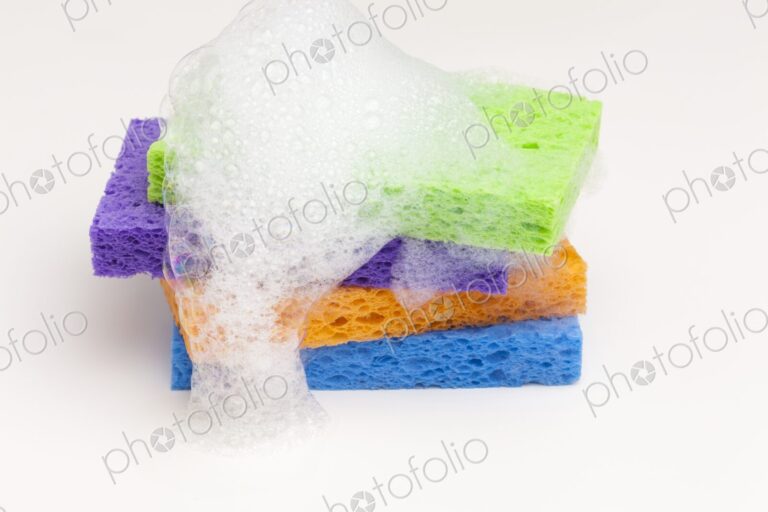 Brightly colored household sponges stacked and covered in foamy soap bubbles against a…
