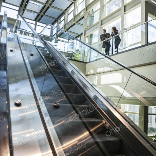Business man and woman talking at the top of an escalator in a large glassed in open… Business man and woman talking at the top of an escalator in a large glassed in open…