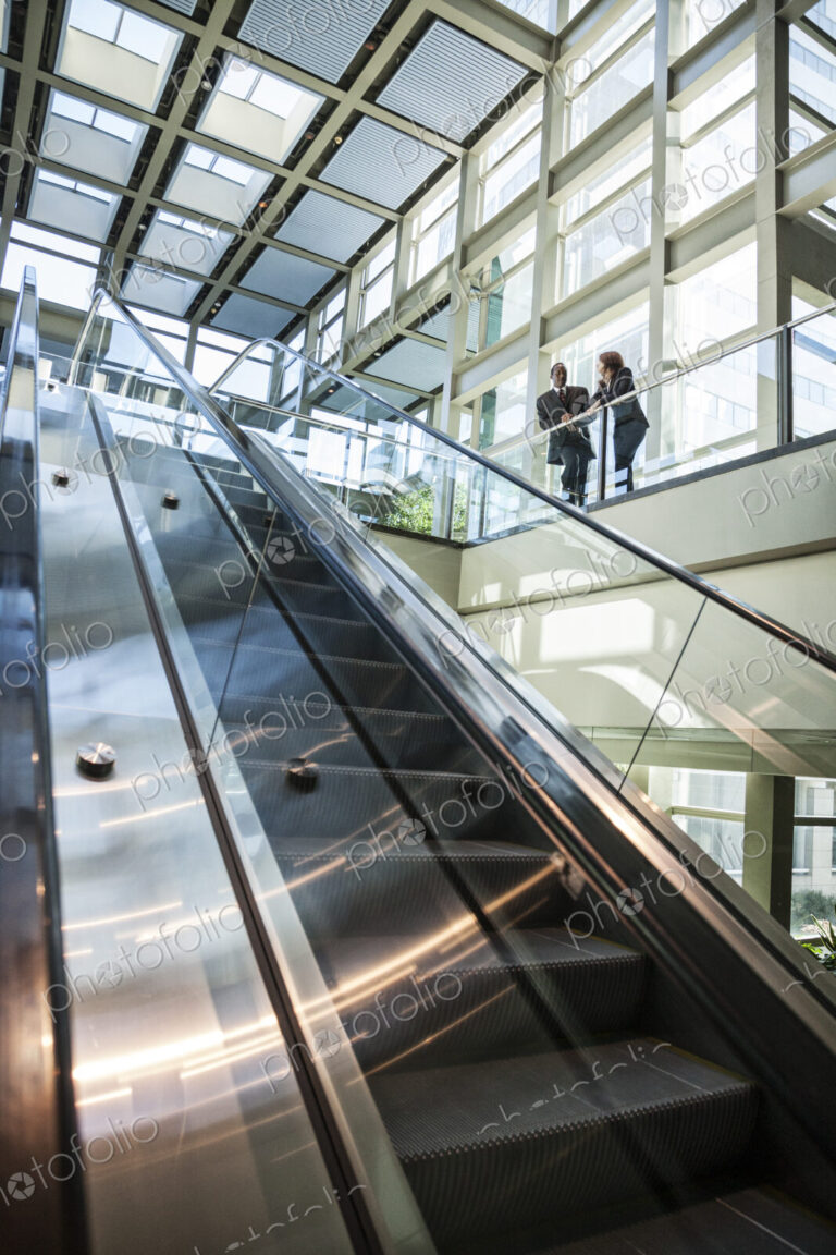 Business man and woman talking at the top of an escalator in a large glassed in open…