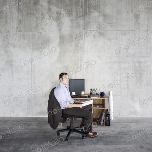 Business man sitting at a small desk in a large empty raw office space.
