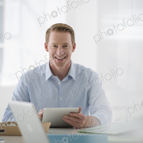 Business. A light airy office environment. A man sitting holding a digital tablet.