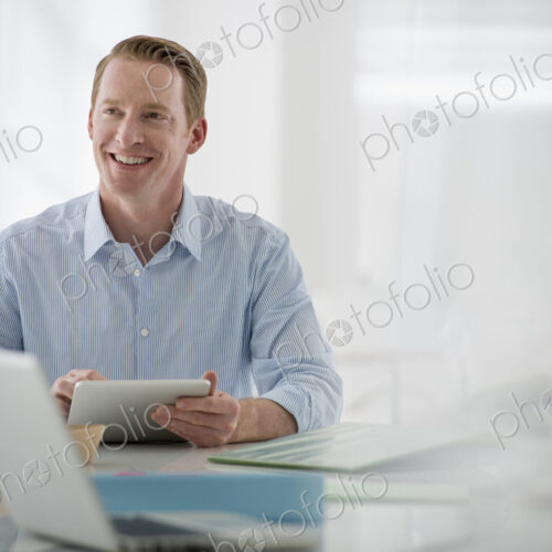 Business. A light airy office environment. A man sitting holding a digital tablet.