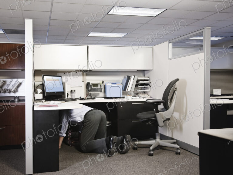 Businessman on his knees in his office cublice trying to connect a plug from a computer.