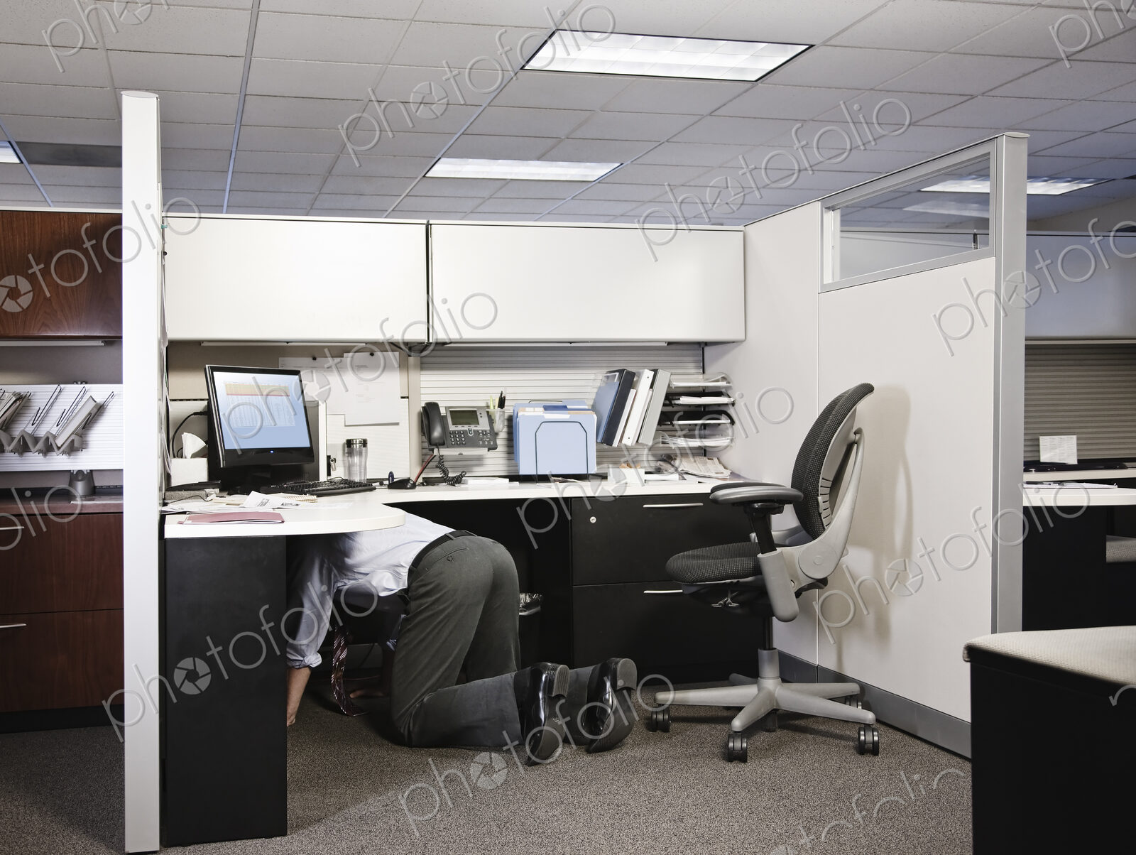Businessman on his knees in his office cublice trying to connect a plug from a computer.