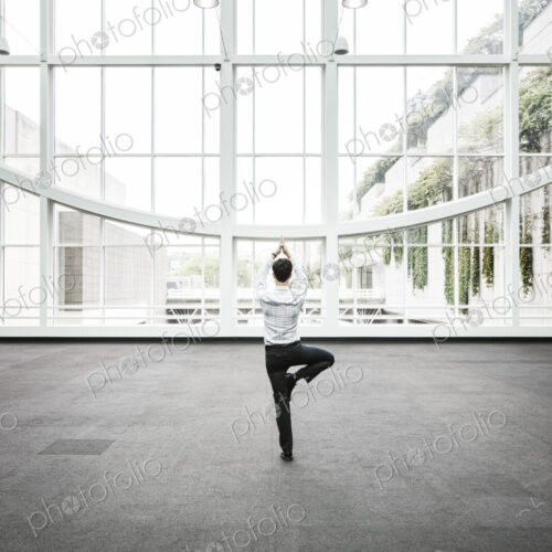 Businessman relaxing doing a yoga pose in a large open glass covered walkway.