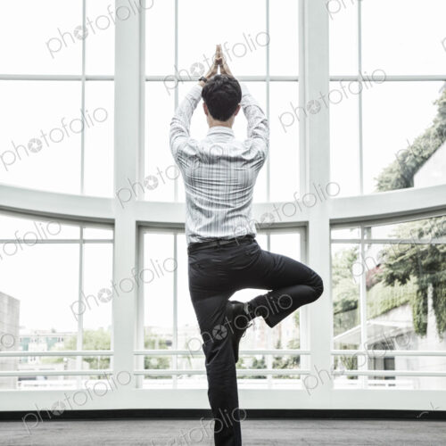 Businessman relaxing doing a yoga pose in a large open glass covered walkway.
