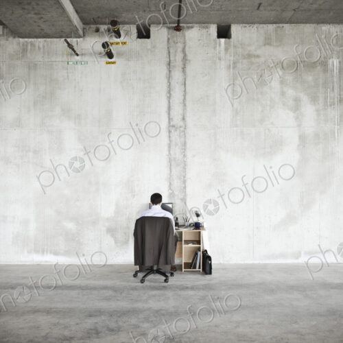 Businessman sitting at a small desk in a large empty raw office space.