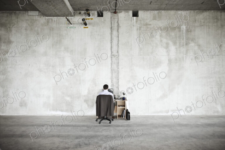 Businessman sitting at a small desk in a large empty raw office space.