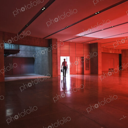Businessman walks through the door of a lobby area that is lit by daylight through red… Businessman walks through the door of a lobby area that is lit by daylight through red…
