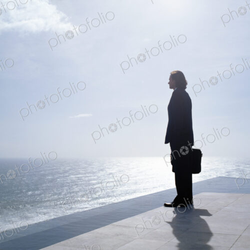 Businessman wearing dark suit holding briefcase, standing outdoors at edge of the ocean.