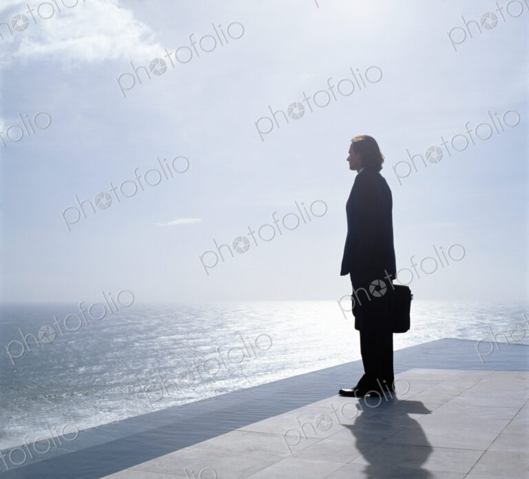 Businessman wearing dark suit holding briefcase, standing outdoors at edge of the ocean.