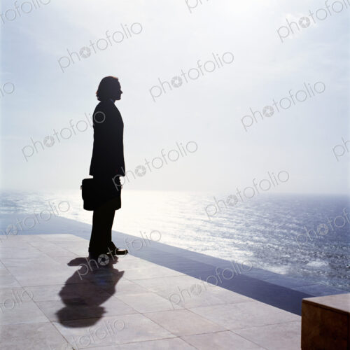 Businessman wearing dark suit holding briefcase, standing outdoors at edge of the ocean.