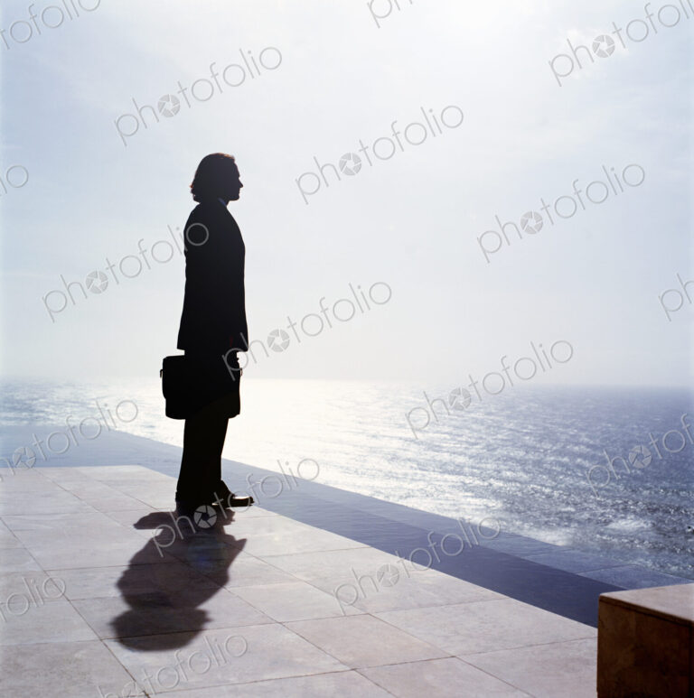 Businessman wearing dark suit holding briefcase, standing outdoors at edge of the ocean.