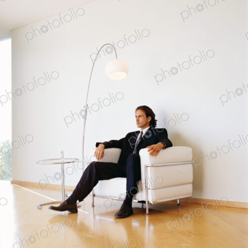 Businessman wearing dark suit sitting indoors on a leather armchair.