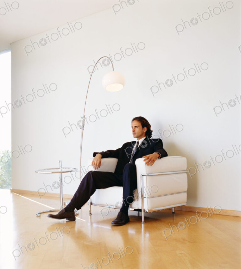 Businessman wearing dark suit sitting indoors on a leather armchair.