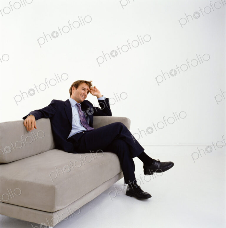 Businessman wearing dark suit sitting indoors on a sofa, hand on head, smiling.