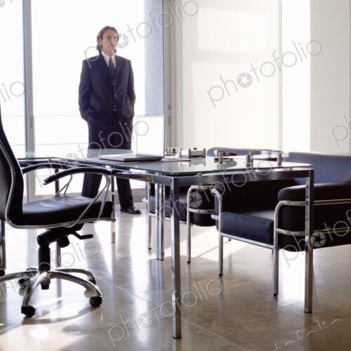 Businessman wearing dark suit standing indoors in an office behind desk with chairs.