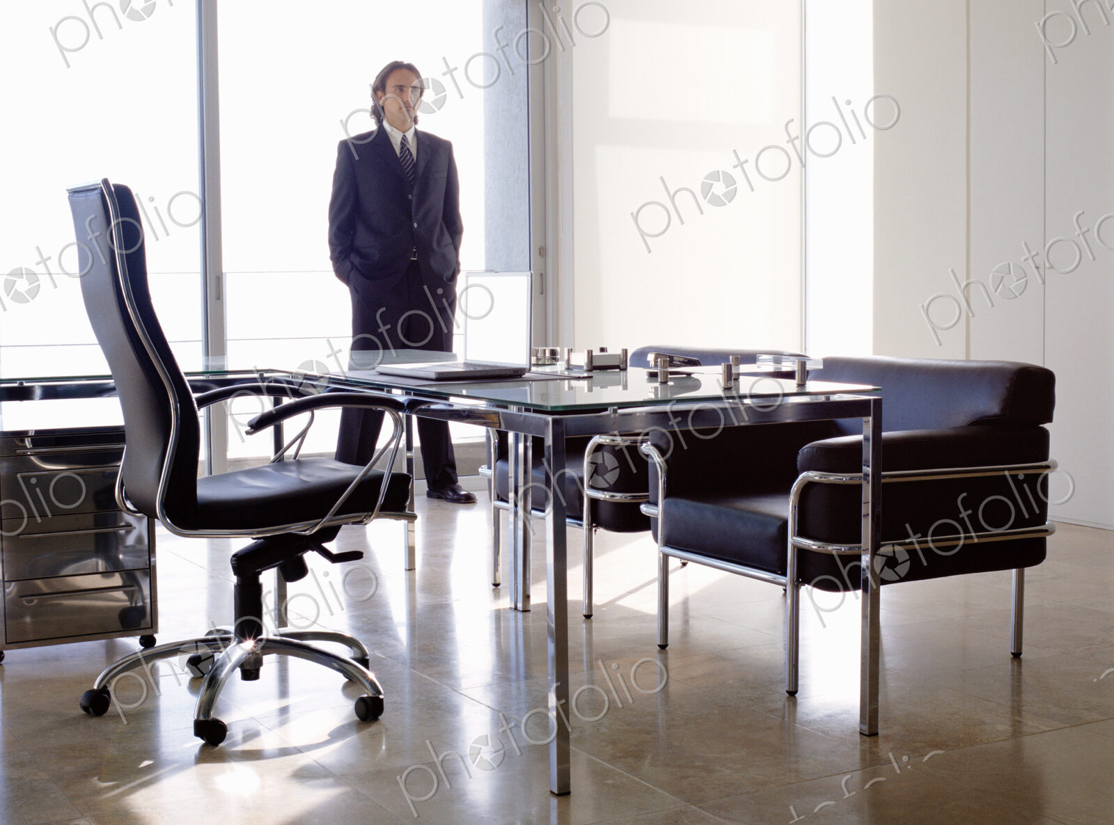 Businessman wearing dark suit standing indoors in an office behind desk with chairs.