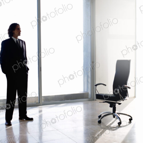 Businessman wearing dark suit standing indoors next to office chair in front of…
