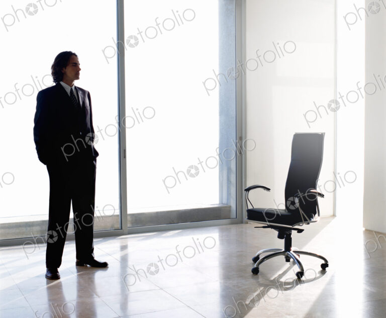 Businessman wearing dark suit standing indoors next to office chair in front of…