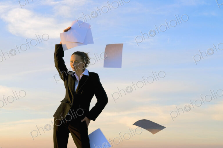 Businesswoman throwing paperwork into the wind with clear sky behind.