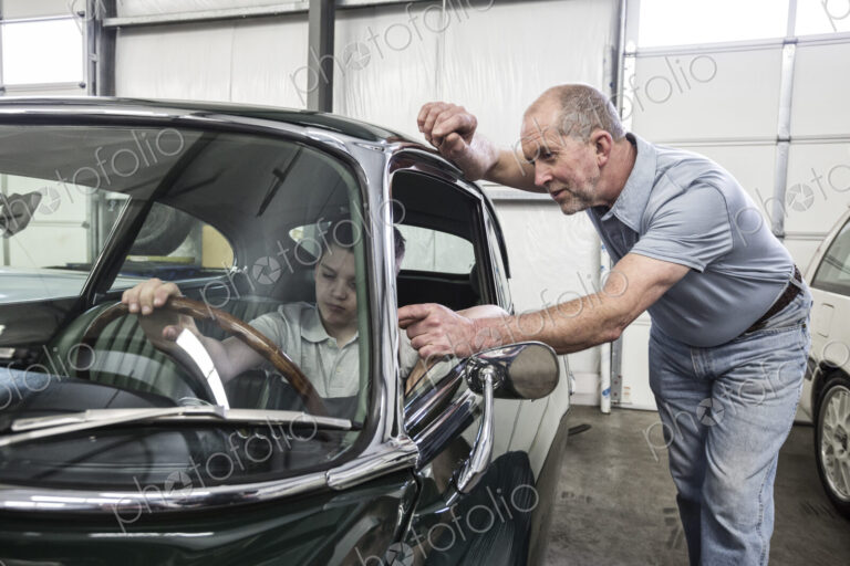 Car mechanic and a small boy sitting in the driving seat of an old classic car.