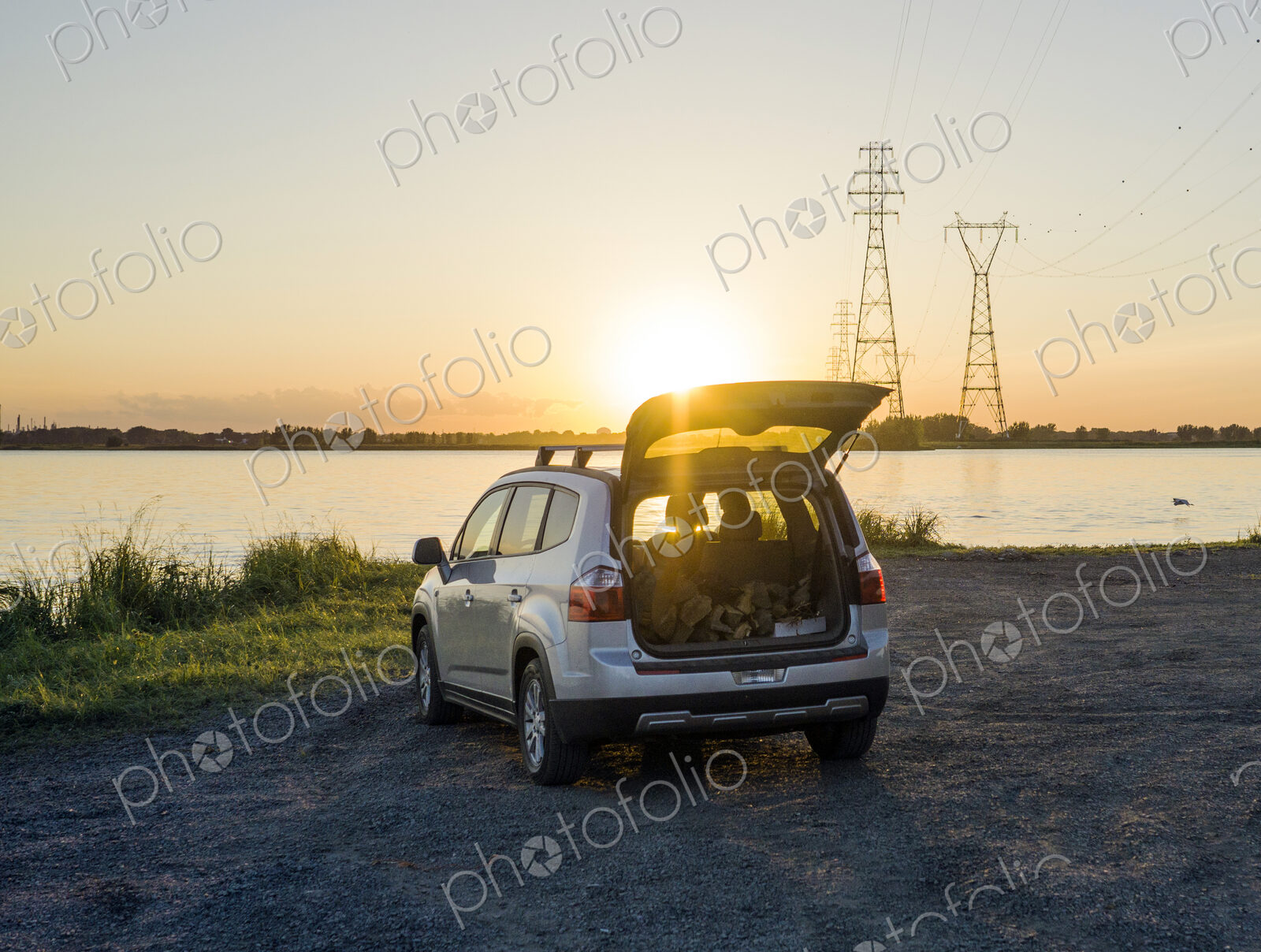 Car with open trunk looking out over a lake or inlet at sunset.