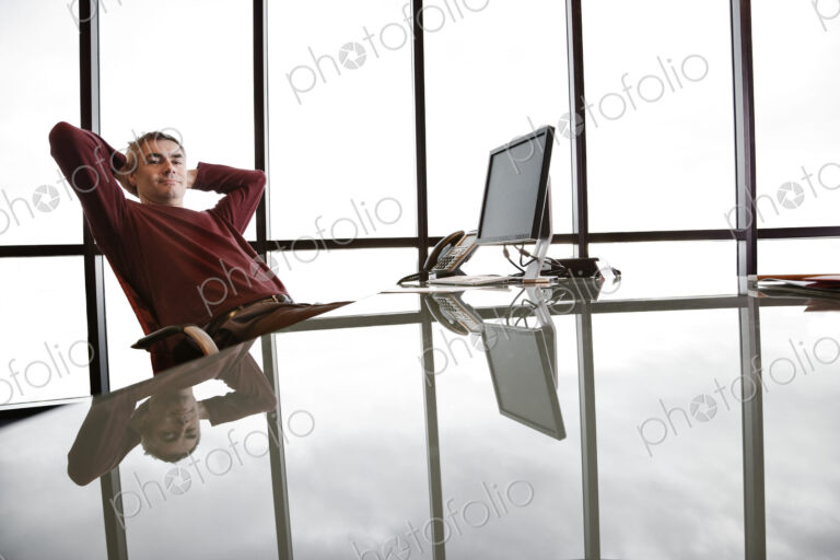 Casually dressed caucasian businessman relaxed at his reflective desk in front of a…
