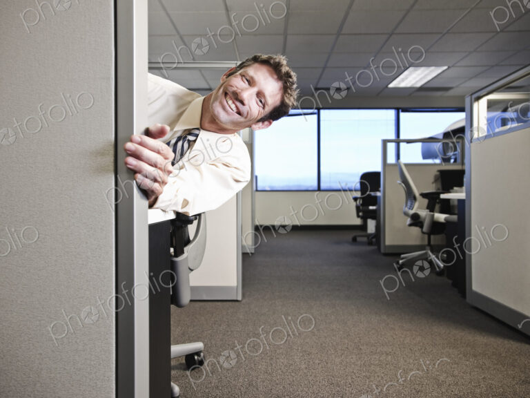 Caucasian businesman smiling and looking around the corner of his office cubicle.