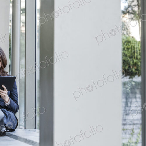 Caucasian business woman using a notebook computer and sitting on a window ledge in a… Caucasian business woman using a notebook computer and sitting on a window ledge in a…