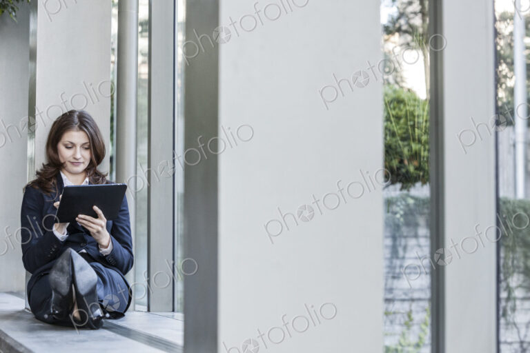 Caucasian business woman using a notebook computer and sitting on a window ledge in a…