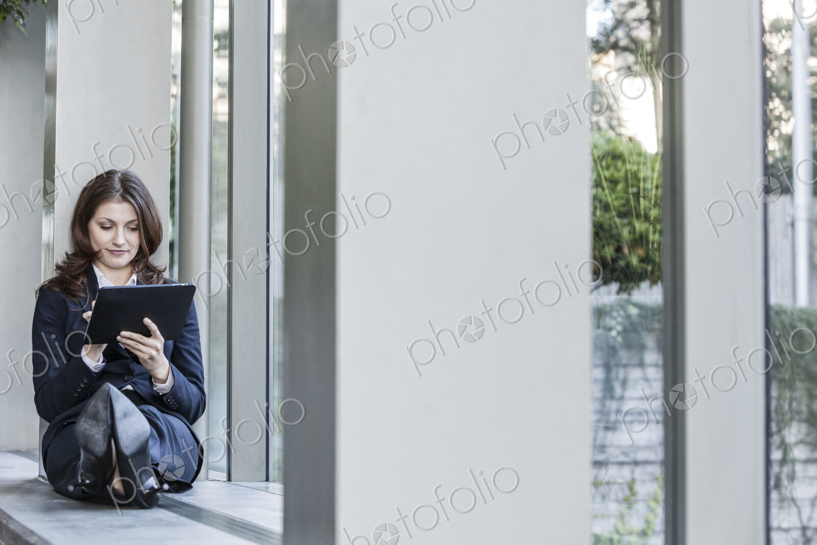 Caucasian business woman using a notebook computer and sitting on a window ledge in a…
